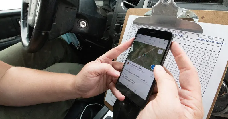 Manos registrando datos de campo en una aplicación del móvil junto a una hoja de registro en papel sujeta a una tablilla